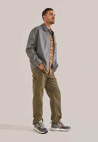 Graue Lederjacke über einem gelben karierten Hemd, olivgrüne Cargo-Hosen und graue Sneakers mit Akzenten. Neutraler Hintergrund.