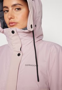 Giacca impermeabile rosa con colletto alto, cappuccio regolabile, bottoni a pressione e il nome del marchio "DIDRIKSONS" sul davanti.