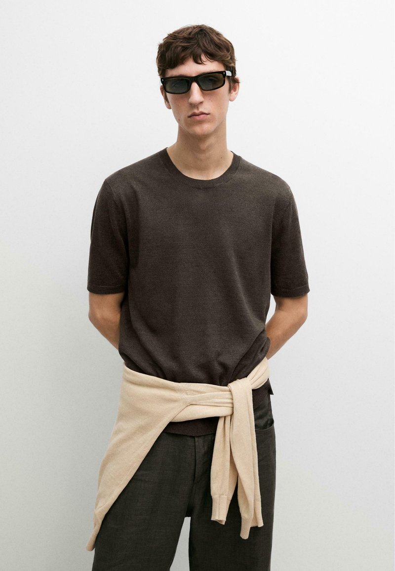 Junger Mann mit kurzen dunklen Haaren trägt schwarze Sonnenbrille, ein dunkelbraunes kurzärmeliges Hemd, dunkle Hosen und einen beigen Pullover, der um seine Taille gebunden ist.