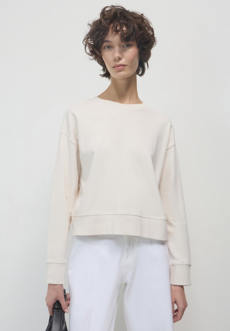 Felpa cropped beige chiaro realizzata in tessuto morbido, con maniche lunghe e scollo rotondo, abbinata a jeans bianchi a vita alta.
