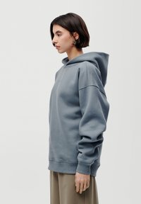 Jeune femme aux cheveux courts et foncés, portant des boucles d'oreilles argentées et un sweat à capuche bleu-gris ample, se tenant de profil contre un fond blanc uni.