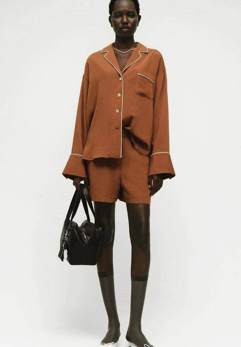 Donna che indossa una camicia button-up di colore ruggine, ampia e comoda, insieme a pantaloni corti coordinati, tiene in mano una borsa nera, con calze scure fino al ginocchio e scarpe bianche.