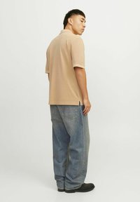 Beige, rövid ujjú póló, oldalsó hasítékokkal, laza szabású kék farmerrel és fekete cipővel viselve. Oldalnézetből rögzítve.