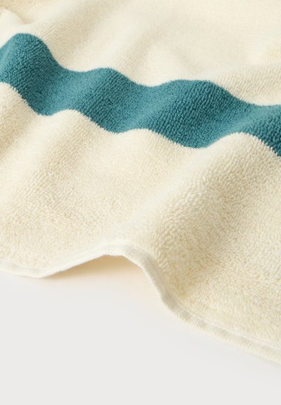 Serviette douce de couleur crème avec une bande texturée en forme de vague bleu sarcelle traversant horizontalement le tissu.