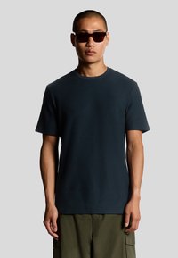 Donkerblauw T-shirt met korte mouwen van gestructureerde stof, met een ronde hals. Draag het met een paar bruine zonnebrillen.
