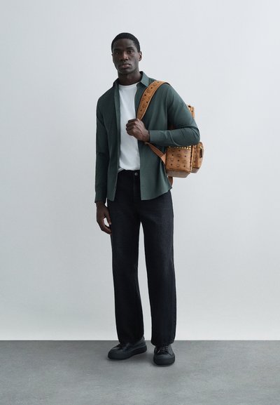 Dark green button-up shirt over a white T-shirt, black wide-leg jeans, black sneakers, and a tan textured backpack with studs.