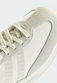 Gros plan sur l'avant d'une basket beige et blanche avec une bordure en daim, des panneaux en tissu et des lacets blancs sur un fond clair.