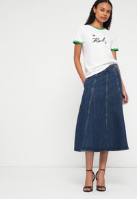 Bijela majica s zelenim obrubom i vezom "Karl", u kombinaciji s dugom plavom traper suknjom s vertikalnim šavovima i lepršavim dizajnom.