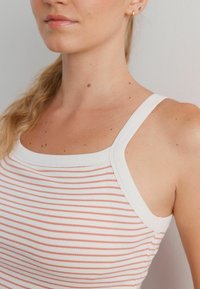 Jeune femme à la peau claire, portant un débardeur rayé blanc et rose, une boucle d'oreille en or et une petite boucle d'oreille bleue.
