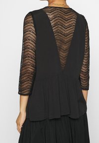 Černá blůza s krajkovými dlouhými rukávy a plisovaným peplum lemem, s hlubokým V-zadním designem a texturovaným zigzagovým vzorem.