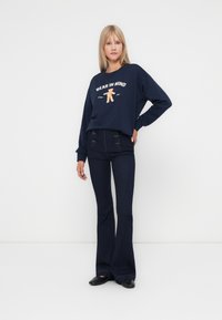 Navy sweatshirt with "BEAR IN MIND" text and bear graphic, paired with flared dark denim jeans. Model wearing black slip-on shoes.