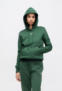 Sweatshirt verde com capuz em material macio, com cordão na gola, bolso frontal e um pequeno logótipo Nike branco no peito. Combina com calças às cores.