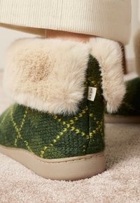 Grøn uldne tøfler med en cremefarvet imiteret pelskant, trekantet gult mønster og en beige sål for komfort. Mærke synligt på siden.