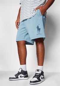 Muž oblečený v světle modrých šortkách Polo Ralph Lauren, pruhovaném tričku, bílých ponožkách a černobílých teniskách Nike, stojící s jednou rukou v kapse.