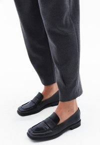 Zwarte leren instappers met een platte zool, voorzien van een verhoogd voorpaneel en stikdetails. Gepaard met grijze, taps toelopende, gemêleerde broek.
