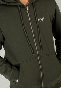 Sudadera con cremallera de color verde oscuro, con un bolsillo frontal, cordones, cremallera de metal y detalle del logo en el pecho. Textura de tela suave.