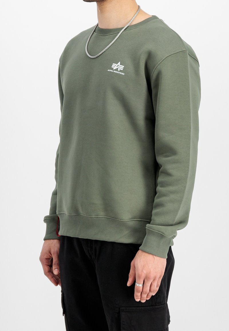 Homme portant un sweat-shirt Alpha Industries vert olive et un pantalon cargo noir, montrant le torse et les bras sur un fond blanc uni.