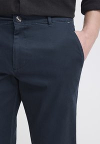 Pravá ruka v kapse tmavě modrých kalhot s viditelným knoflíkem a poutkem na pásek, osoba má na sobě černý dlouhý rukáv.