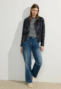 Giubbotto in pelle nera, camicia a righe, jeans blu a gamba larga e sneakers chiare, posizionati contro un muro neutro. Outfit casual con texture sovrapposte.