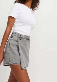 Mini gonna in denim grigio con design avvolgente, dotata di chiusura a bottone e cuciture a vista. Indossata con una t-shirt bianca aderente.