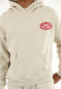 Sudadera gris hecha de una tela suave, con un bolsillo frontal y dobladillo acanalado. Logo bordado centrado en rojo.
