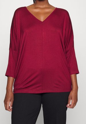 Femme portant un haut ample à manches longues, col en V profond de couleur rouge foncé, et un pantalon noir, debout devant un arrière-plan clair et uni.