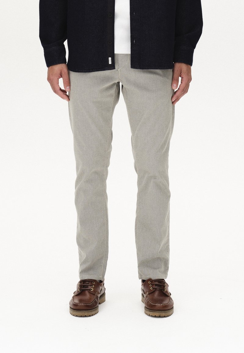 Homme portant un pantalon gris clair, des chaussures en cuir marron foncé, une chemise blanche et une veste noire, debout devant un fond blanc uni.