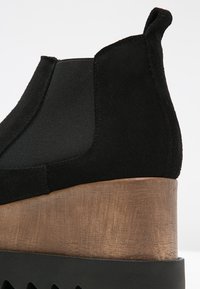 Bottine en suède noir avec des panneaux latéraux élastiques et un talon en bois en coin. Le talon présente une surface texturée avec un léger éclat.