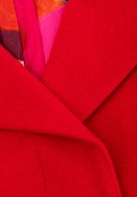 Rode wollen jas met een reverskraag, gemaakt van een gestructureerde stof. Een kleurrijke zijden sjaal in roze en oranje piept onder de kraag vandaan.