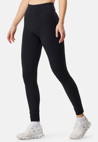 Leggings neri a vita alta con una texture liscia e un design aderente, abbinati a sneakers sportive bianche con una suola distintiva.