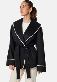 Czarny płaszcz z szerokim klapami, białym falbaniastym wykończeniem, długimi rękawami i paskiem do wiązania, wykonany z teksturowanej tkaniny.