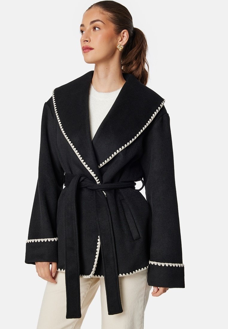 Czarny płaszcz z szerokim klapami, białym falbaniastym wykończeniem, długimi rękawami i paskiem do wiązania, wykonany z teksturowanej tkaniny.