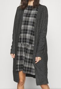 Dlouhý šedý cardigan vyrobený z měkké pleteniny, otevřený vpředu, s žebrovanými manžetami, nošený přes černobílé kostkované šaty s rozšířenou sukní.