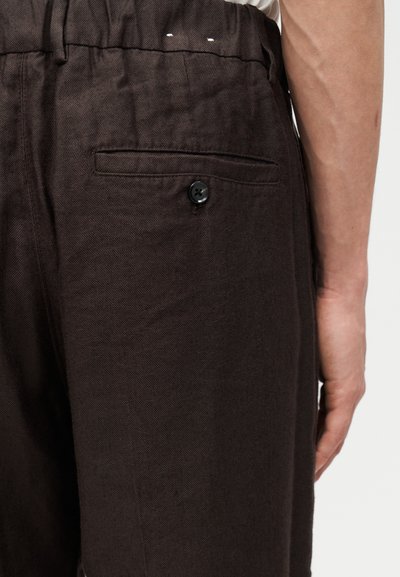 Κοντινό πλάνο ενός ατόμου που φοράει σκούρο καφέ παντελόνι με πίσω τσέπη που ασφαλίζει με μαύρο κουμπί, εμφανίζοντας μέρος του δεξιού χεριού.