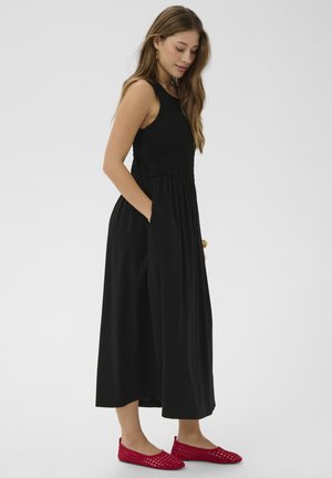 Frau in einem ärmellosen schwarzen Midikleid mit Taschen und roten gewebten Ballerinas, seitlich stehend vor einem schlichten hellen Hintergrund.