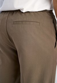 Braune, elastische Bundhosen mit glatter Textur und einer einzelnen Gesäßtasche. Der Stoff scheint leicht zu sein und hat einen dezenten Glanz.