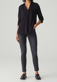 Blouse noire à manches longues avec col, offrant une coupe décontractée et une encoche au niveau du col, associée à un jean slim gris foncé et des mocassins noirs.