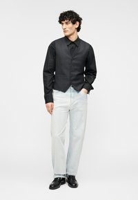 Μαύρο πουκάμισο με κουμπιά κάτω από ένα ραμμένο γιλέκο σκούρου γκρι χρώματος, τζιν παντελόνι σε ανοιχτό μπλε με ίσια γραμμή και μαύρα παπούτσια. Λιτή σχεδίαση με απαλό ύφασμα.