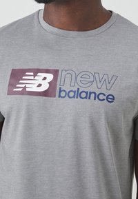 Szara koszulka z krótkim rękawem, wykonana z miękkiego materiału, z dużym bordowym i niebieskim logo "new balance" z przodu. Z klasycznym dekoltem.