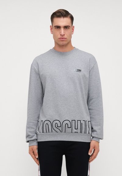Homme portant un sweatshirt gris Moschino avec du texte noir près de l'ourlet et sur la poitrine, associé à un pantalon noir avec des bandes latérales.