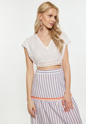 Lyshåret kvinde iført en hvid cropped top med bølget kant og et stribet skørt med en orange blomsterkant, ser til siden.