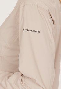 Lys beige langærmet skjorte med en glat tekstur, sømdetaljer og et sort "ENDURANCE" logo på venstre ærme.