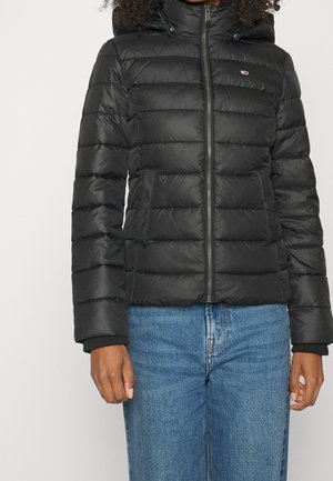 Person iført en sort quiltet dunjakke med lynlås foran og blå jeans, stående mod en ensfarvet baggrund.