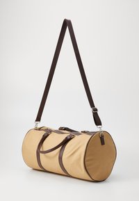 Sac de sport en toile beige, avec des accents en cuir marron, deux poignées et une longue sangle réglable. Forme arrondie, fermeture éclair.