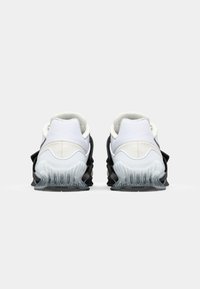 Vue arrière de chaussures de sport présentant un dessus blanc, un panneau en maille texturé et une semelle noire ondulée avec des accents transparents.
