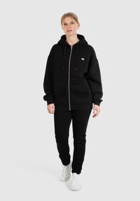 Schwarzer Reißverschluss-Hoodie mit großen Fronttaschen, dickem Stoff und Kordelzugkapuze. Kombiniert mit passenden schwarzen Hosen und weißen Turnschuhen.