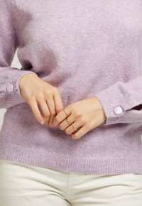 Pull en maille violet avec une finition texturée et des boutons lavande ronds aux poignets. Deux anneaux en or sur les mains, déposés sur un tissu léger.