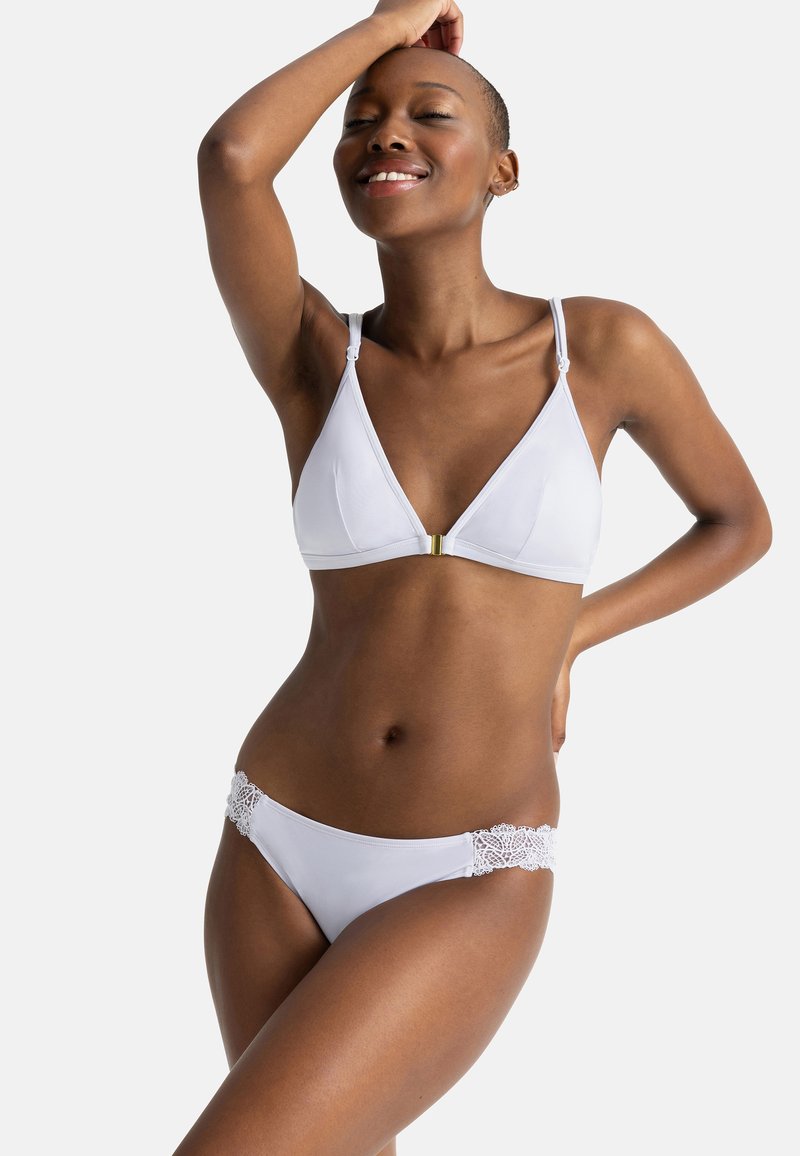 Baltas bikini, turintis trikampį viršų su auksiniu akcentu ir apačią su nėriniu ant klubų, pagamintas iš lygaus audinio.