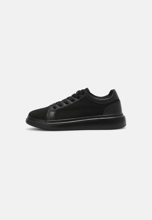 Czarny niski sneaker z fakturowaną cholewką z tkaniny, sznurowaniem i grubą gumową podeszwą na jasnym, jednolitym tle.