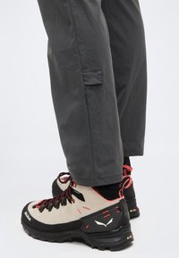 Μπεζ και μαύρα μποτάκια πεζοπορίας με κόκκινες λεπτομέρειες, φορεμένα με σκούρο γκρι παντελόνι πάνω σε λευκό φόντο.